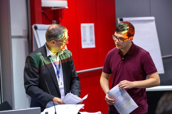 Photo capturing interaction between an older man on the left and a younger man on the right who is holding a piece of paper