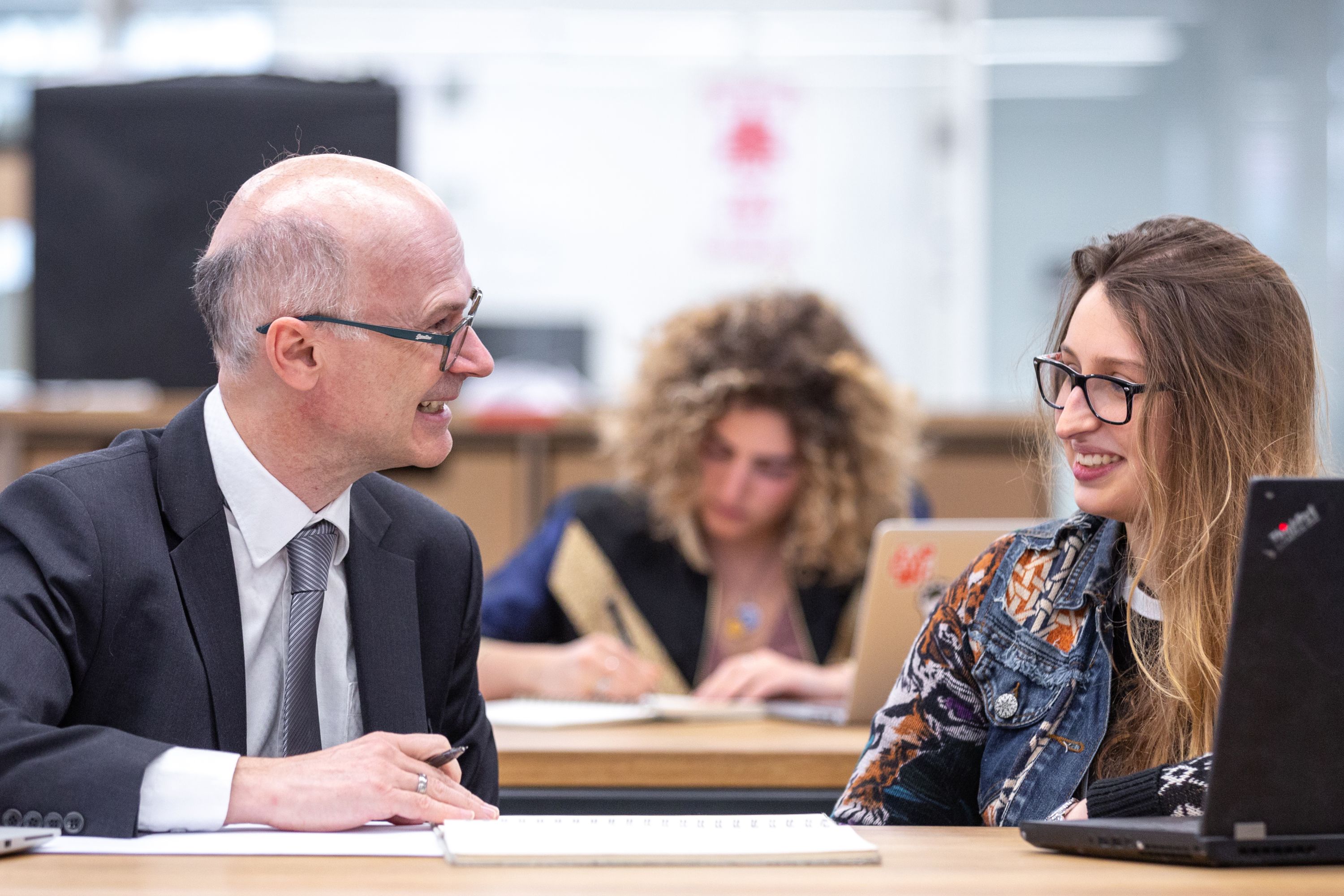 Two people chatting at a desk, and a student working behind