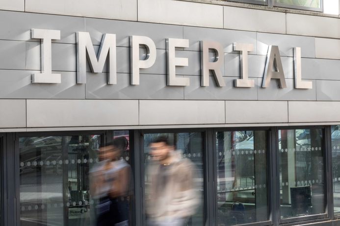 A long-exposure photo of Imperial's branding on a building, with blurred students walking past on the pavement